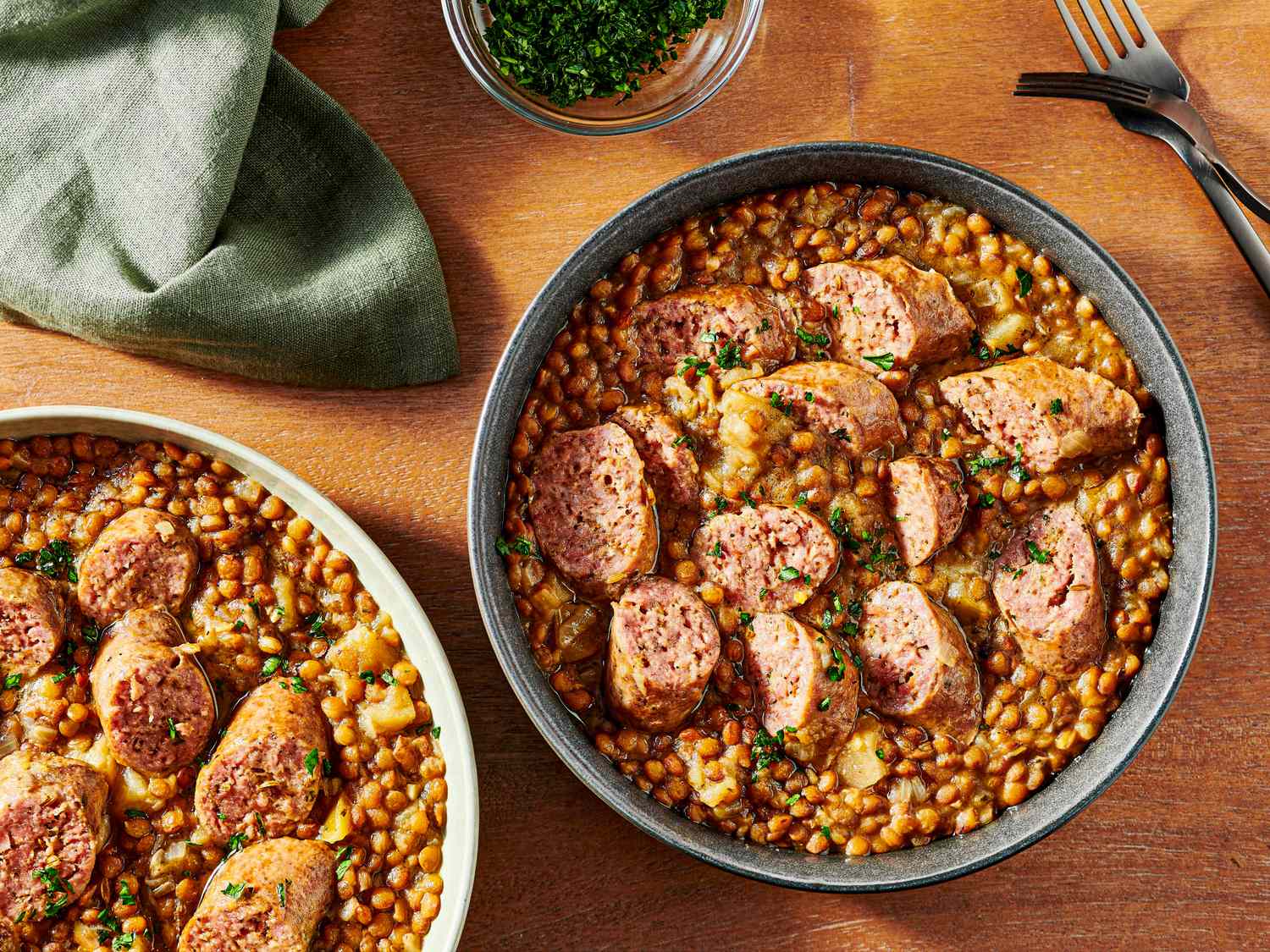 2 bowls of pressure cooked lentils and sausage on a wooden tabletop, with a soft turquoise napkin, two forks, and small bowl of parsley for garnish to the sides. 