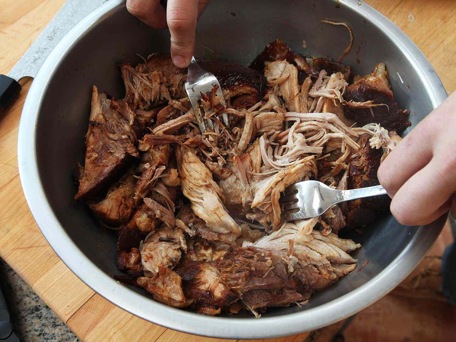 Pork being shredded and pulled apart in a bowl with two forks.