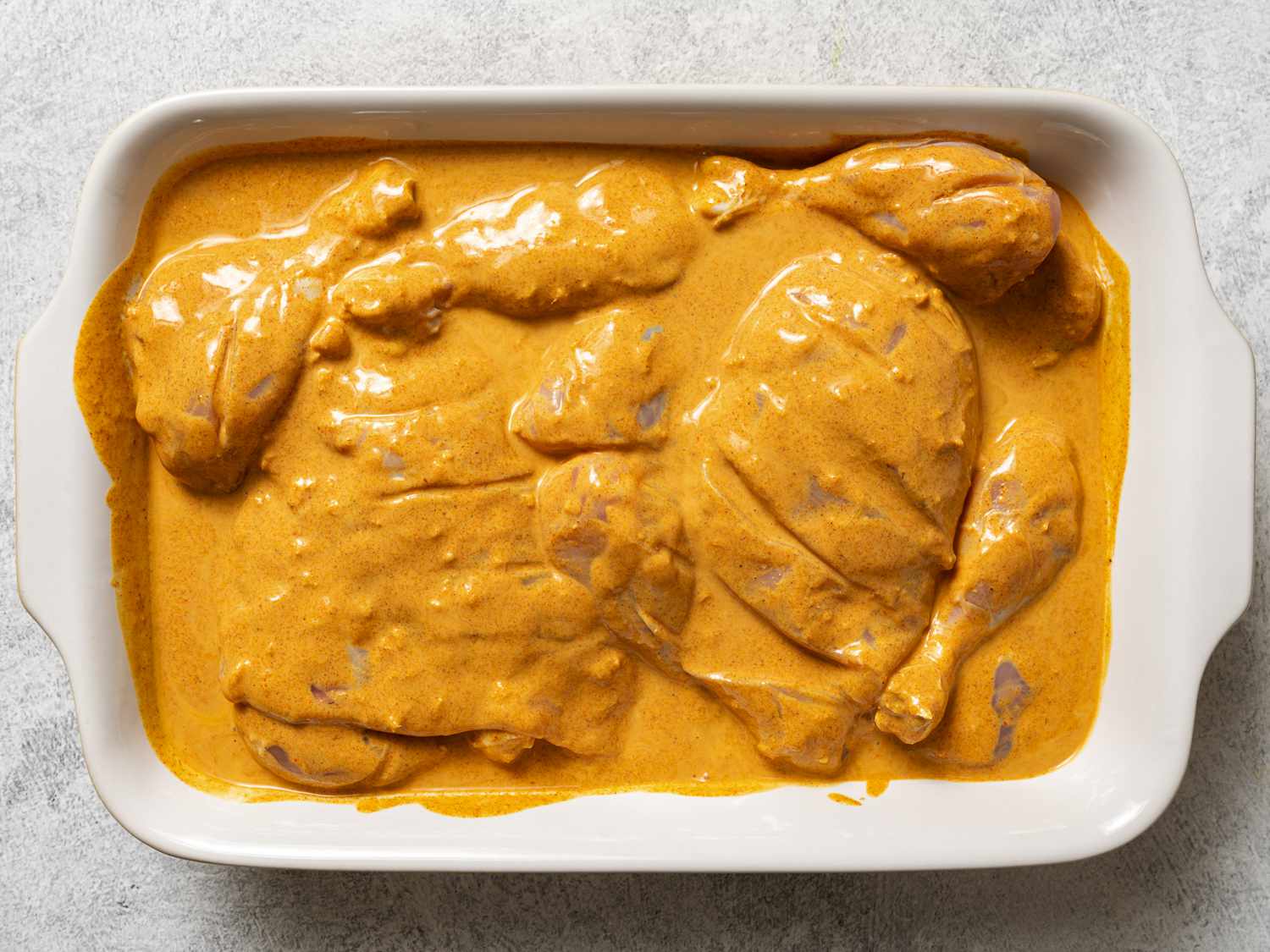 The chicken pieces and marinade inside of a rectangular baking dish. The chicken is thoroughly coated in the marinade.