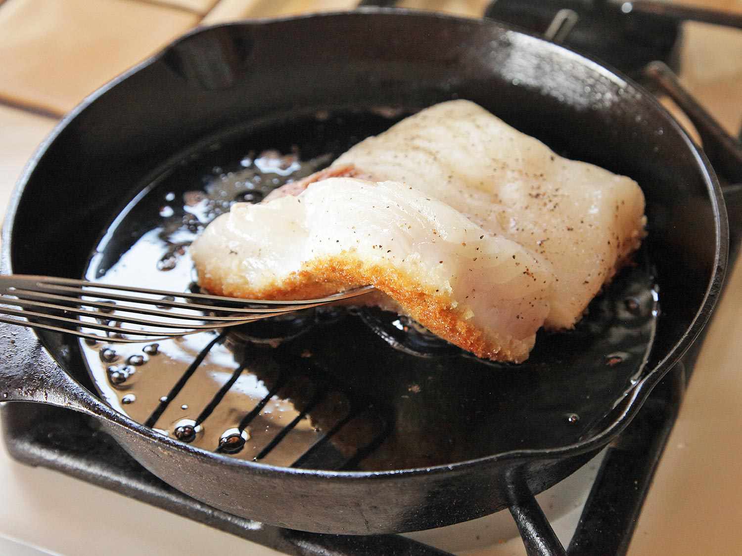 A slotted fish turner lifts one of the frying fish fillets to reveal a golden brown breadcrumb crust.