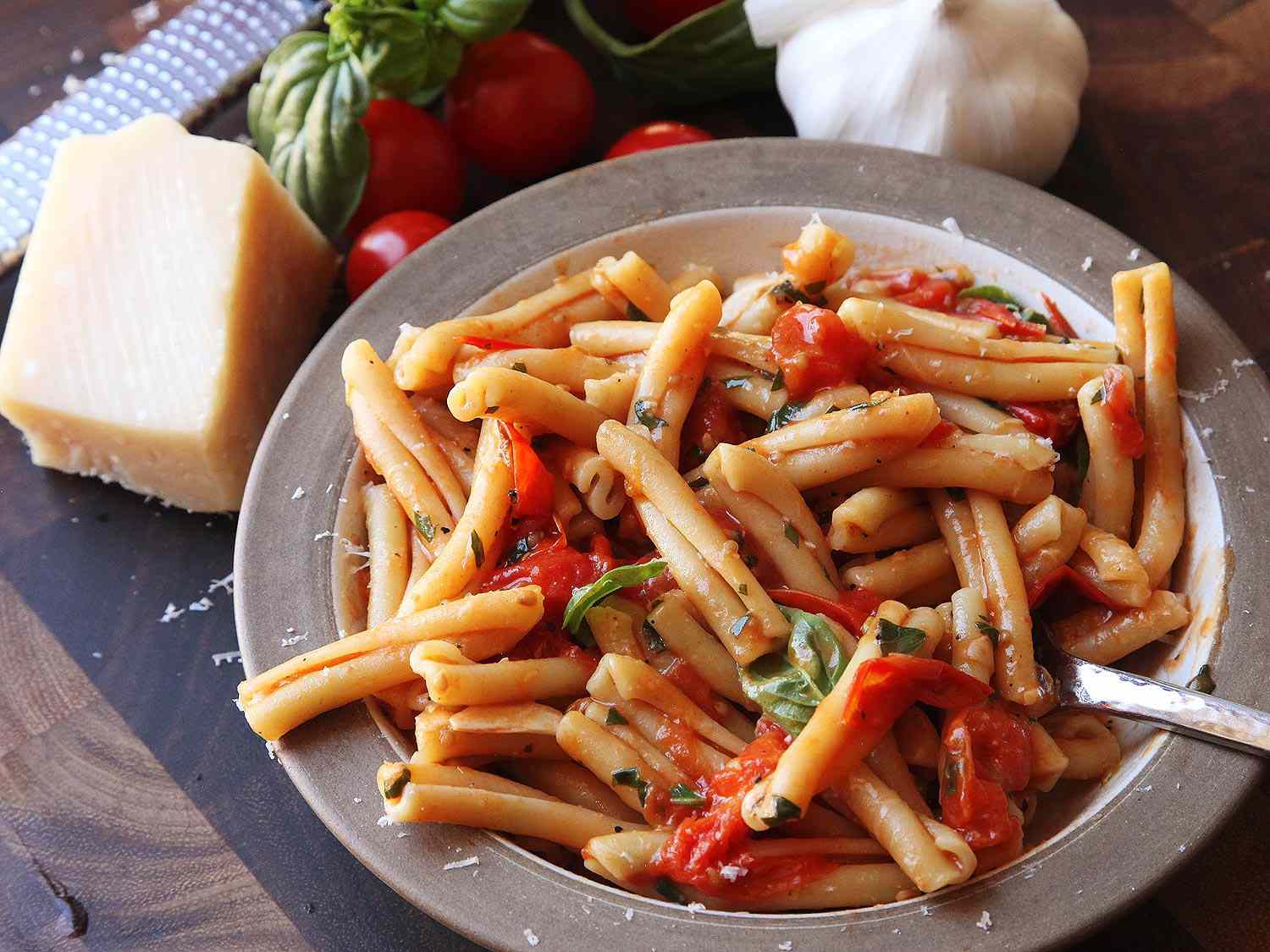 Finished dish of pasta with blistered cherry tomato sauce, next to a block of cheese, basil sprig, garlic, and tomatoes.