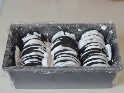 A loaf pan lined with plastic wrap, with whipped cream and three rows of cookies to make up the inside of the icebox cake.