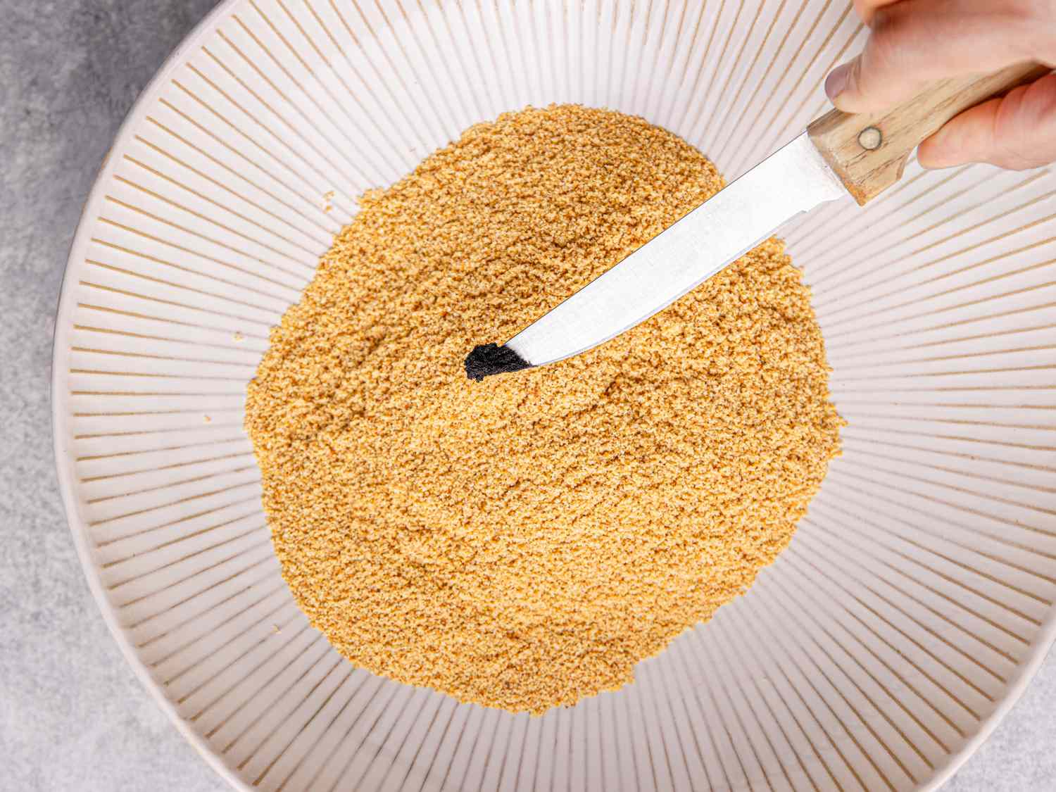 A hand holding a vanilla bean pod over a bowl of powdered ingredients with a knife scraping seeds from the pod into the mixture