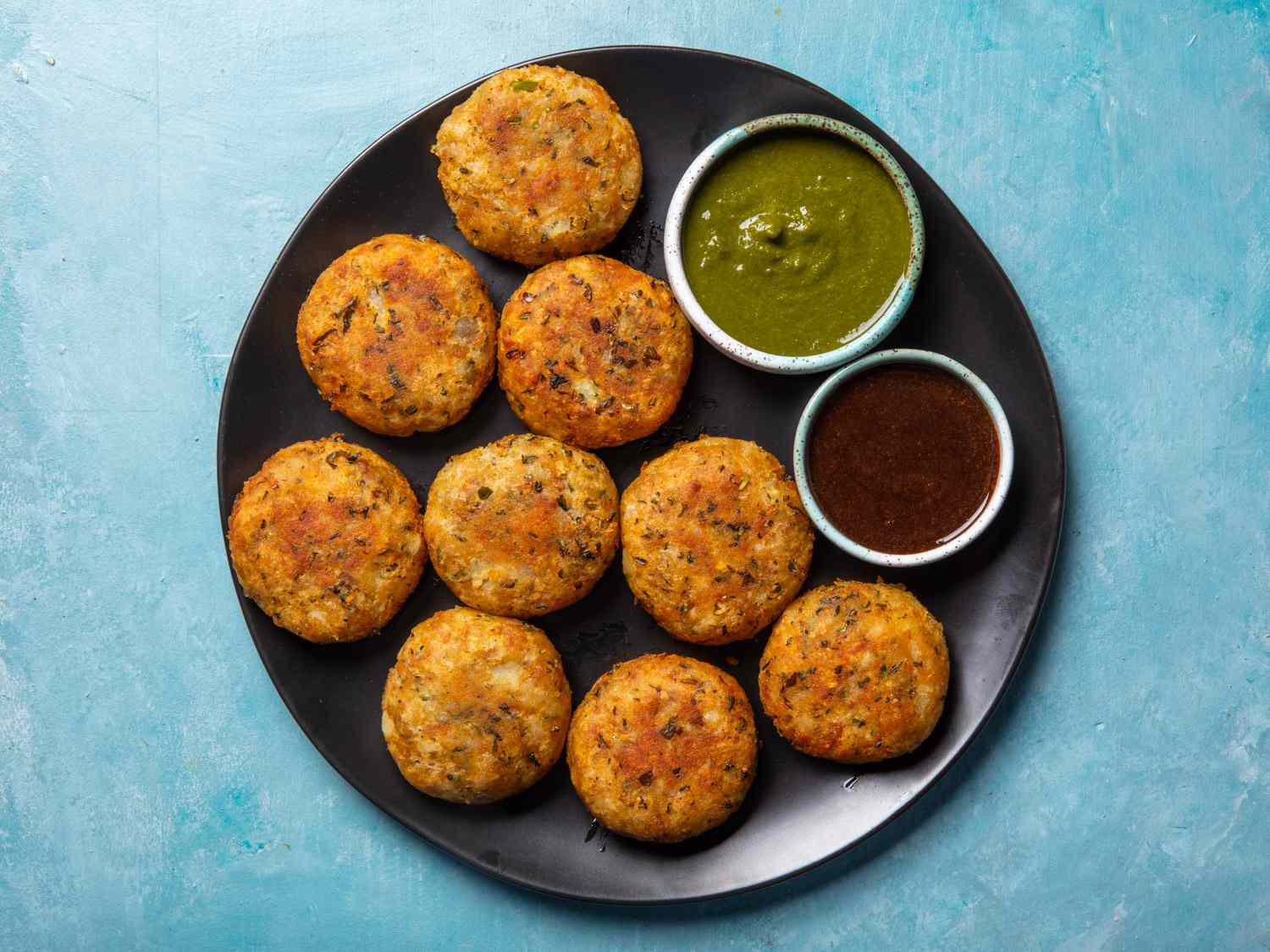 Overhead view of aloo tikki on a plate with dippers of mint and tamarind chutneys alongside