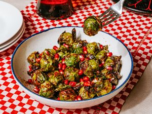 A bowl of airfried Brussels sprouts garnished with pomegranate seeds with a fork picking up a piece placed on a checkered tablecloth