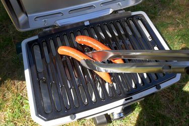 a person using a pair of grill tongs to flip a hot dog on a portable gas grill