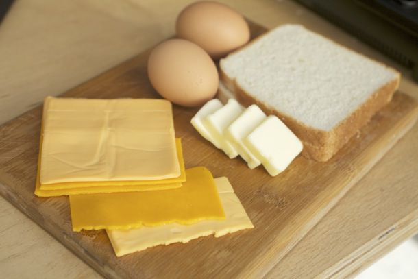 Several slices of cheese (American and cheddar), two eggs, four pats of butter, and two slices of bread on a wooden cutting board.