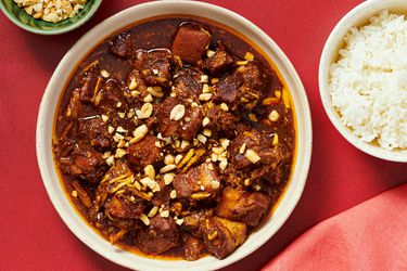 Overhead of stew in a bowl with small dish of peanuts in bowl to the side with red napkin and red background 