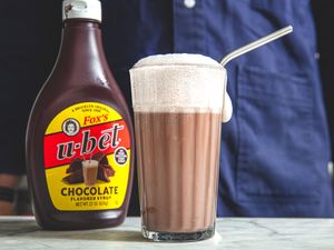 A glass of chocolate egg cream with a metal straw next to a bottle of Ubet chocolate syrup