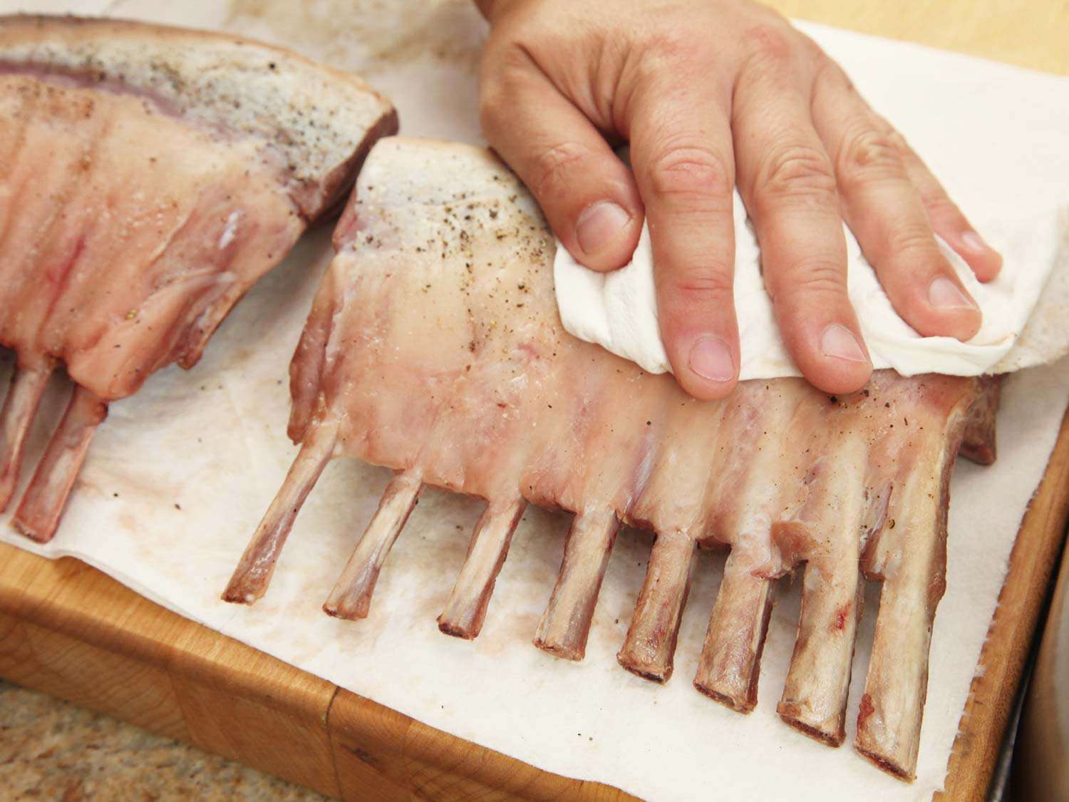 Hands patting two sous vide lamb racks dry with paper towels.