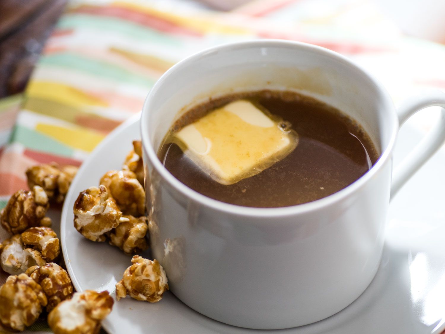 Closeup of Hot Caramel-Popcorn Bourbon Apple Cider, served in a mug with a pat of butter floating on the surface of the cocktail. Caramel corn is piled nearby.