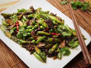 Stir-fried spring vegetables with black olives and Sichuan peppercorns, served on a square platter.