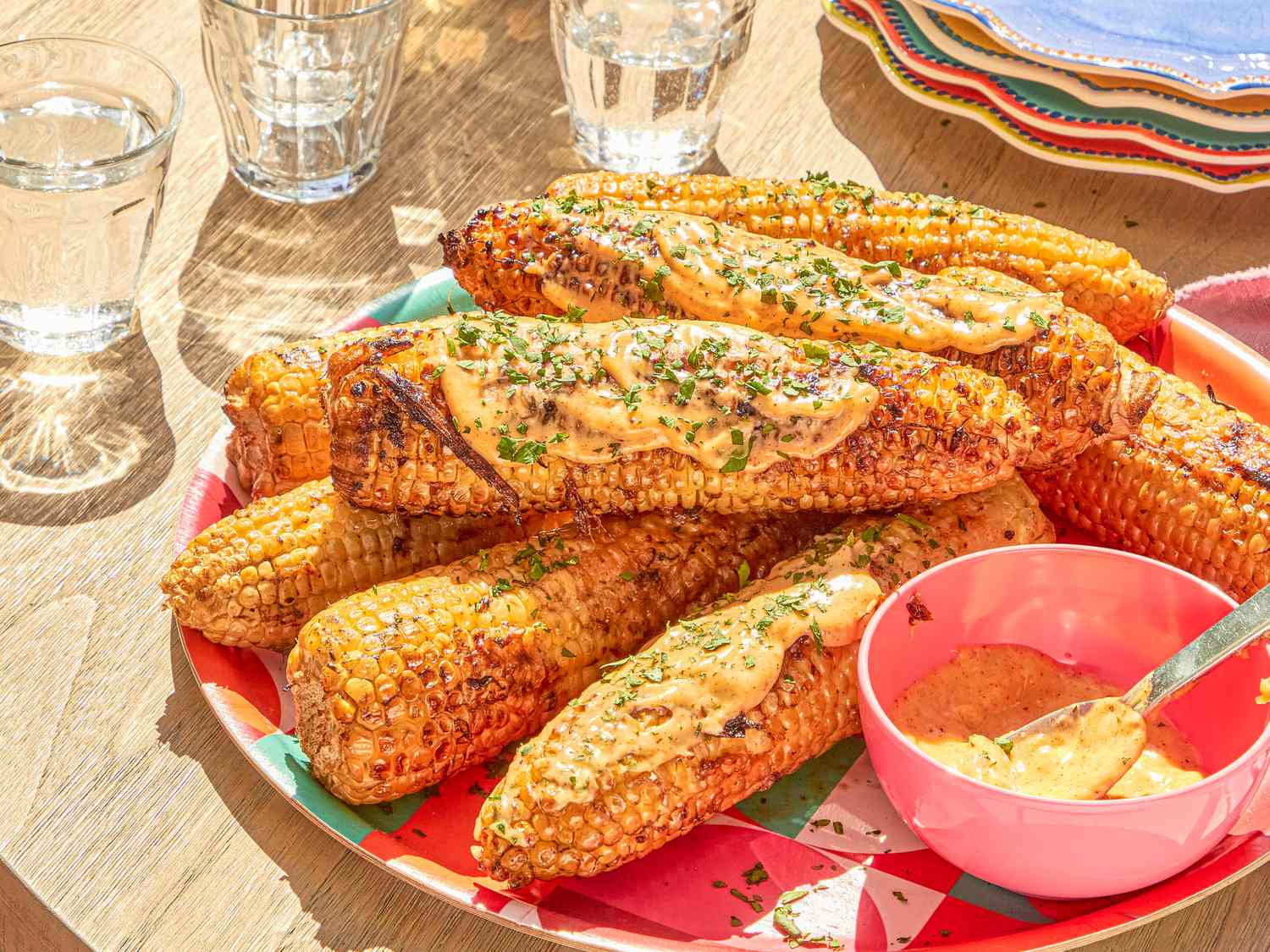Plated grilled corn cobs topped with sauce and herbs alongside a small bowl of sauce