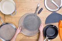 a variety of splatter screens on a wooden surface with someone placing a screen over a pan