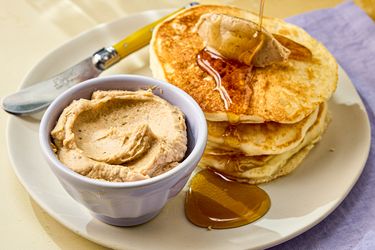 A plate of pancakes served with a small bowl of spreadable butter and honey with a yellowhandled knife nearby