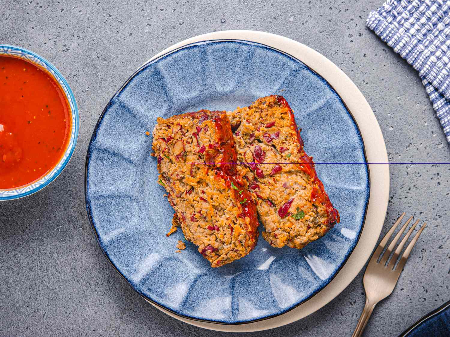 Two slices of meatloaf on a plate with a side of sauce and cutlery placed nearby