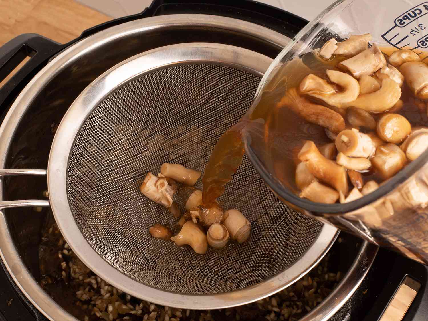Mushroom stock being poured into the pressure cooker through a fine mesh sieve, with the sieve catching the solid pieces of mushroom.