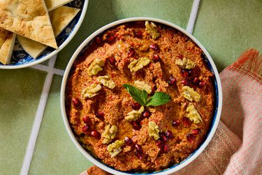 A bowl of muhammara garnished with walnuts, pomegranate arils, and a mint leaf, next to pita bread on a green tile surface