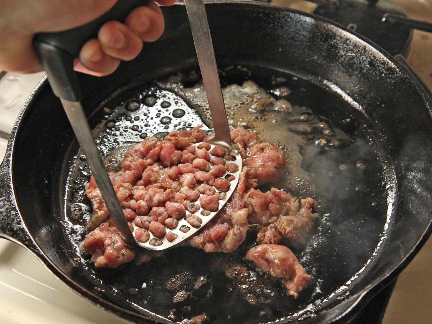 Mashing sausage meat with a potato masher in a cast iron skillet. 
