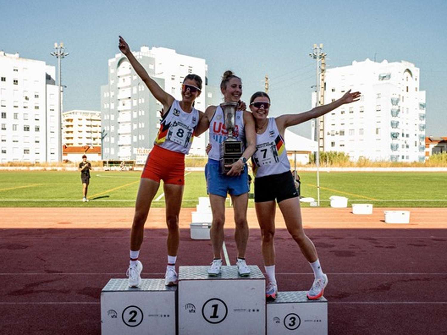 Three athletes standing on a podium the center individual holding a trophy with others celebrating on either side outdoor sports setting