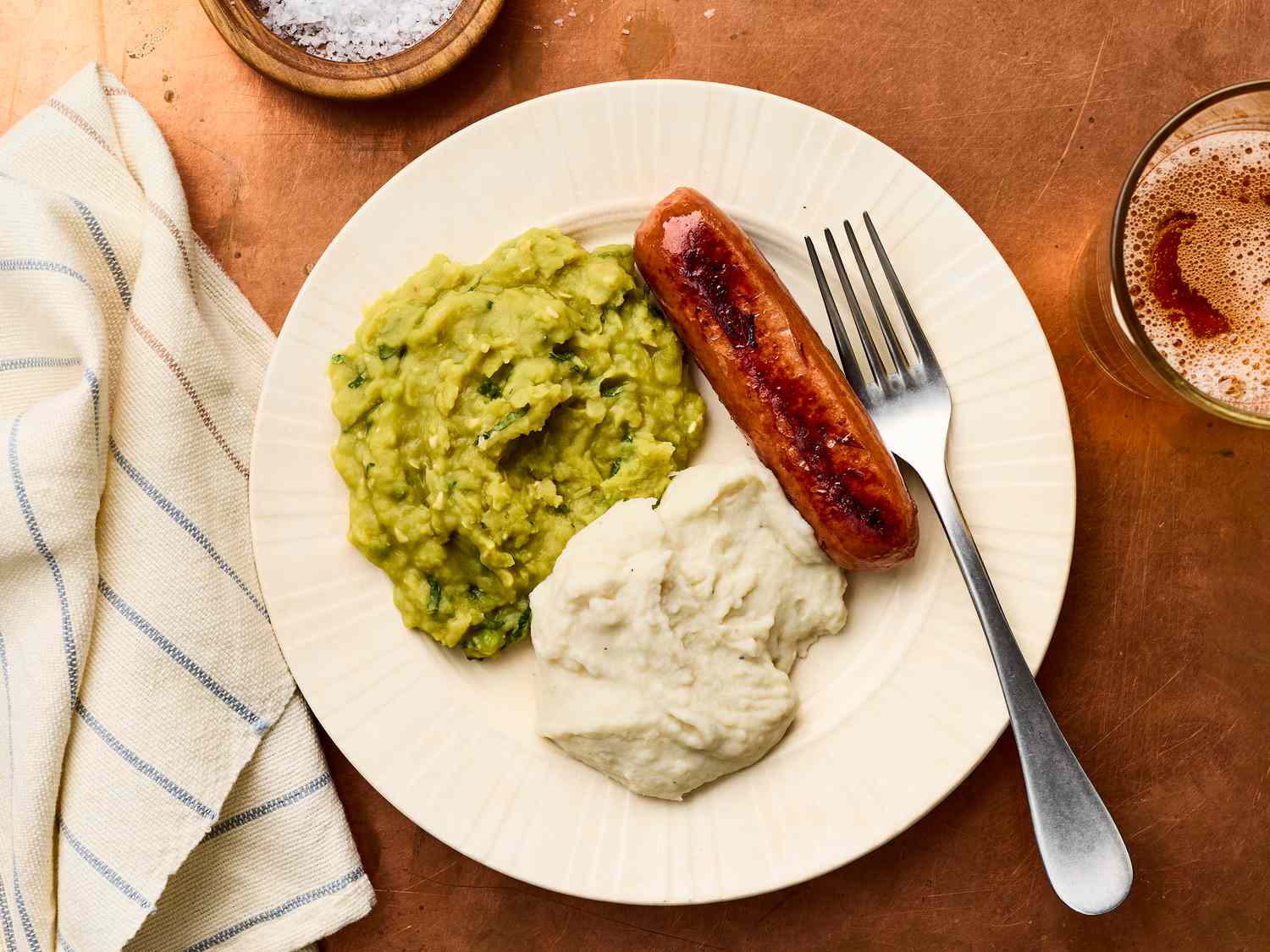 A plate with mashed peas, mashed potatoes, a sausage, and a fork resting on the edge