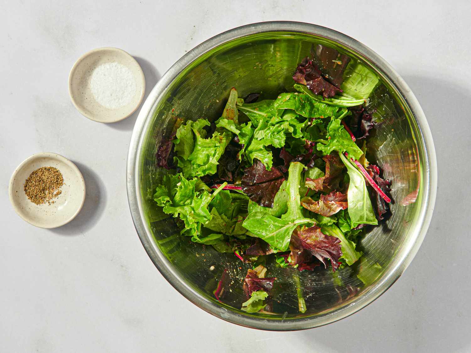 A metal bowl of mixed greens next to small dishes of salt and pepper