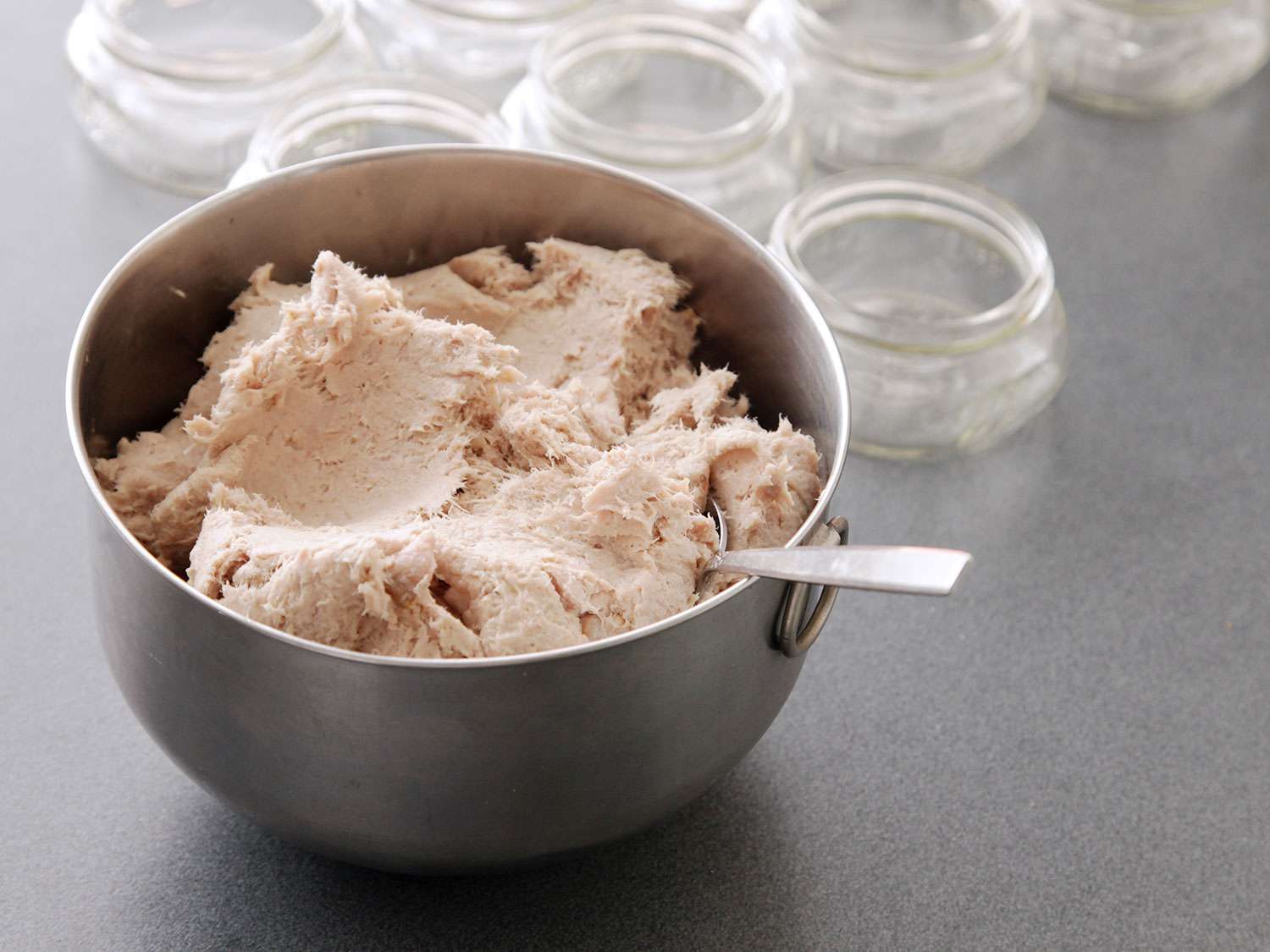 A small metal mixing bowl filled with pork rillettes. Wide-mouth quarter-pint jars are arrayed in the background.