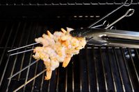 A closeup look at tongs flipping two metal skewers that have shrimp threaded onto them