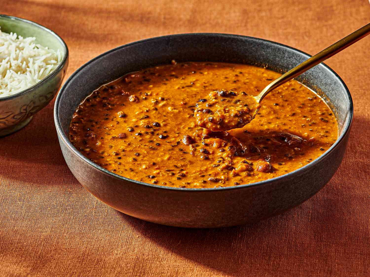 Large bowl of dal with a spoonful of some, smaller bowl of white rice on the side, on top of a burnt orange textile 