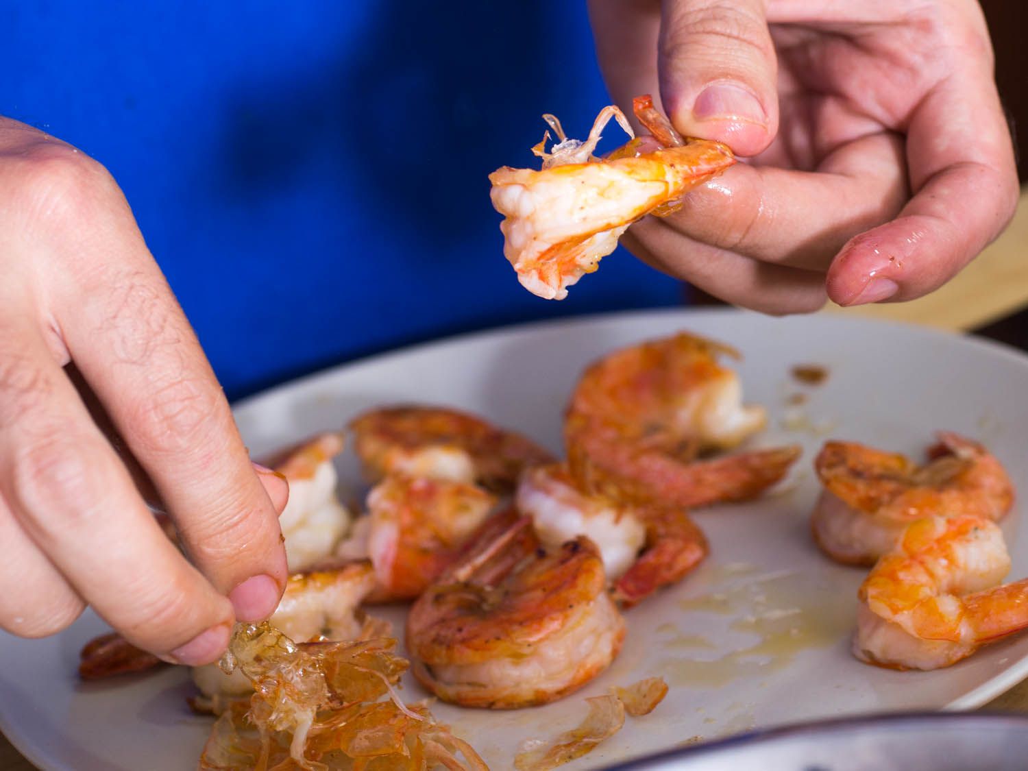 Hands peeling cooked shrimp on a white plate.