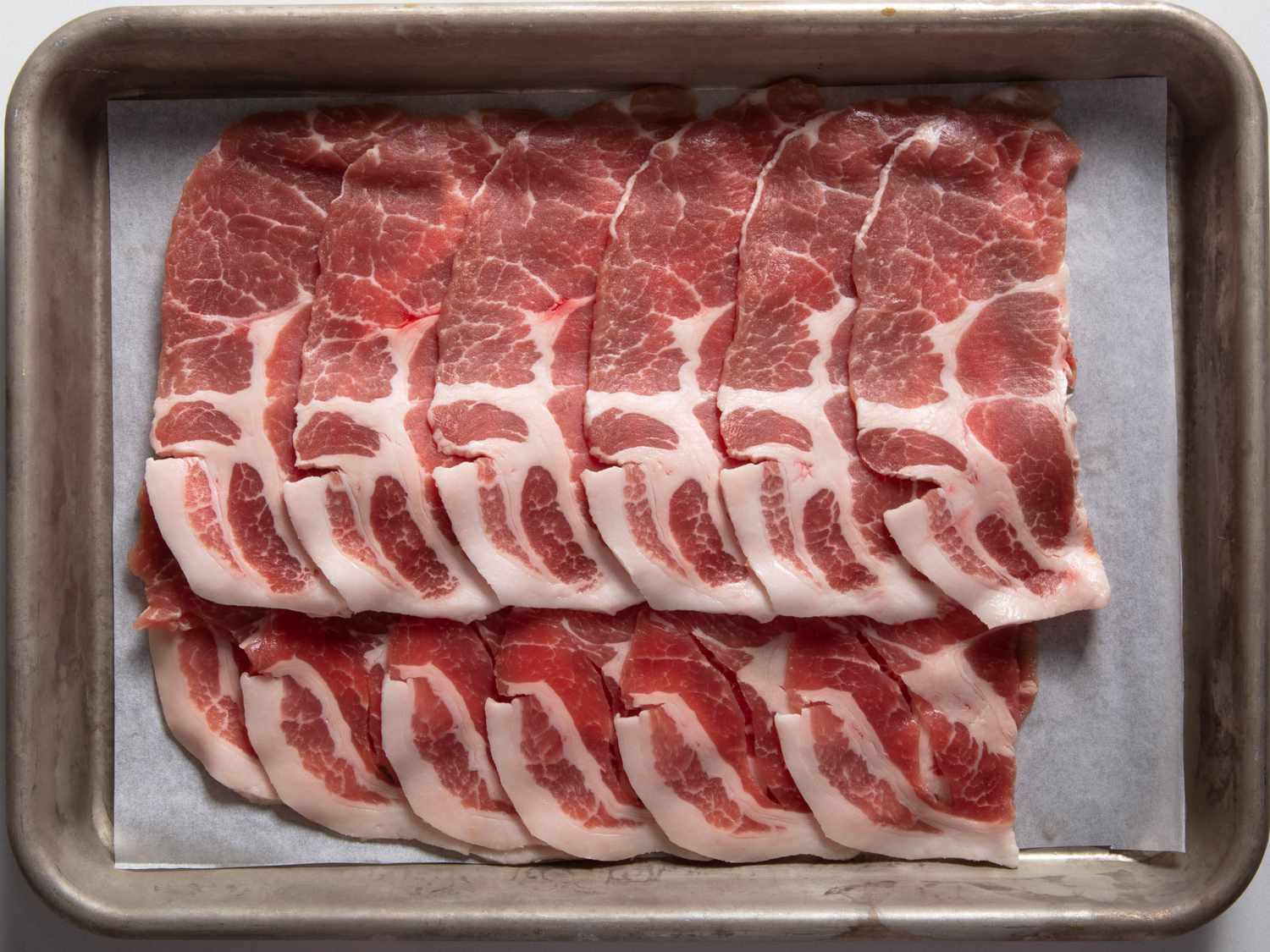 Thin slices of raw pork shoulder fanned out on a rimmed baking sheet