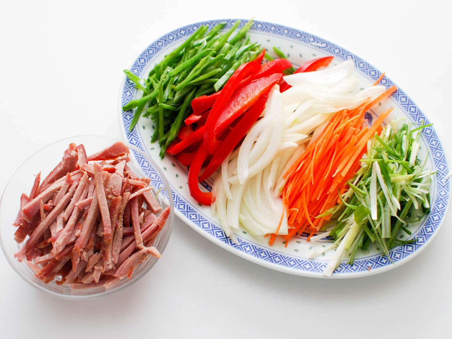 Thin sliced snow peas, red bell peppers, onion, carrot, and scallions on a platter next to a small bowl of thinly sliced char siu.