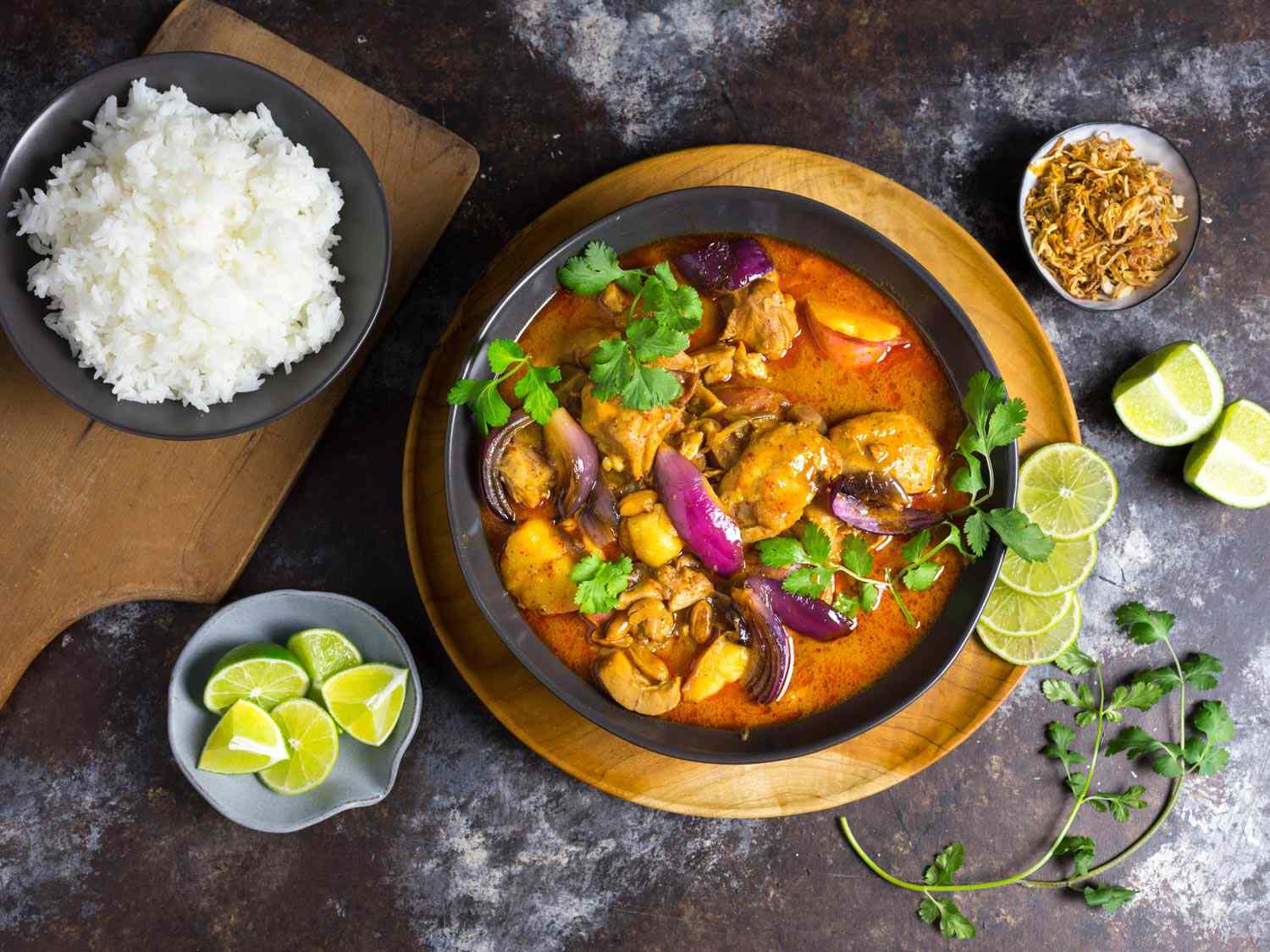 Overhead view of chicken massaman curry with wheat beer and potatoes, served in a gray bowl alongside rice, crispy shallots, and lime wedges.