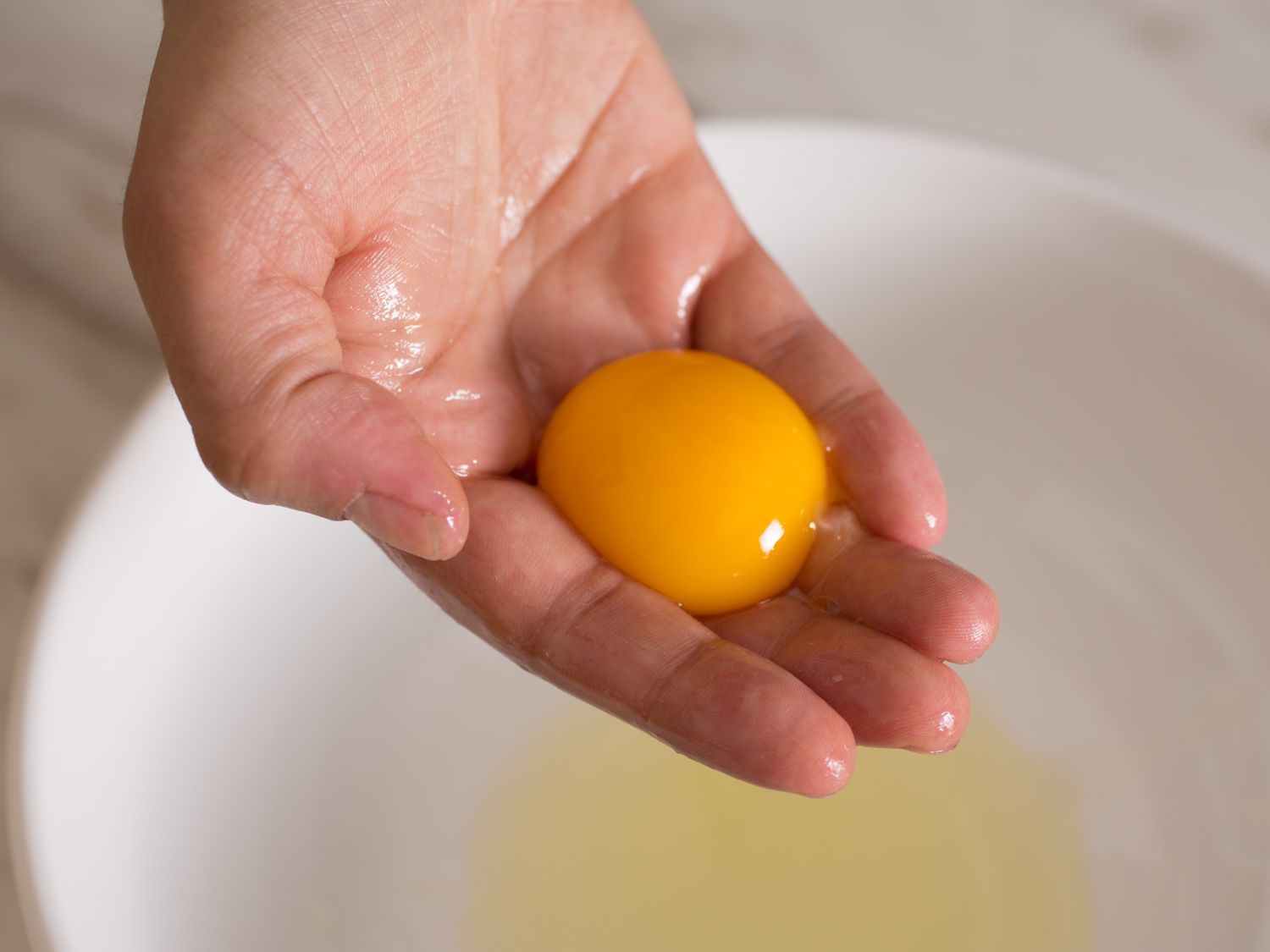 A hand cradling an egg yolk with egg white in a white bowl in the background.