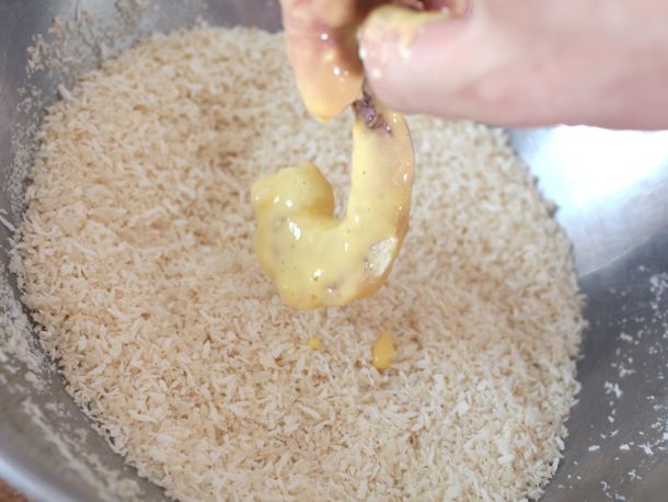 A hand dipping a shrimp covered in batter into a stainless steel bowl holding shreds of dried coconut mixed with panko breadcrumbs.