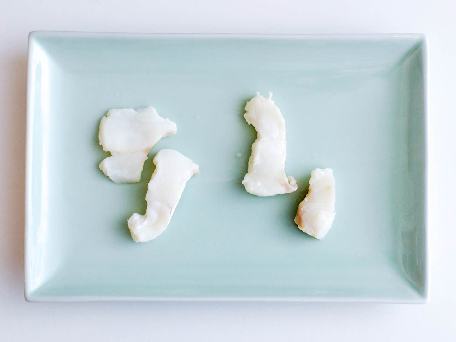 Two pieces of water-blanched cod on a plate next to two pieces of oil-blanched cod.