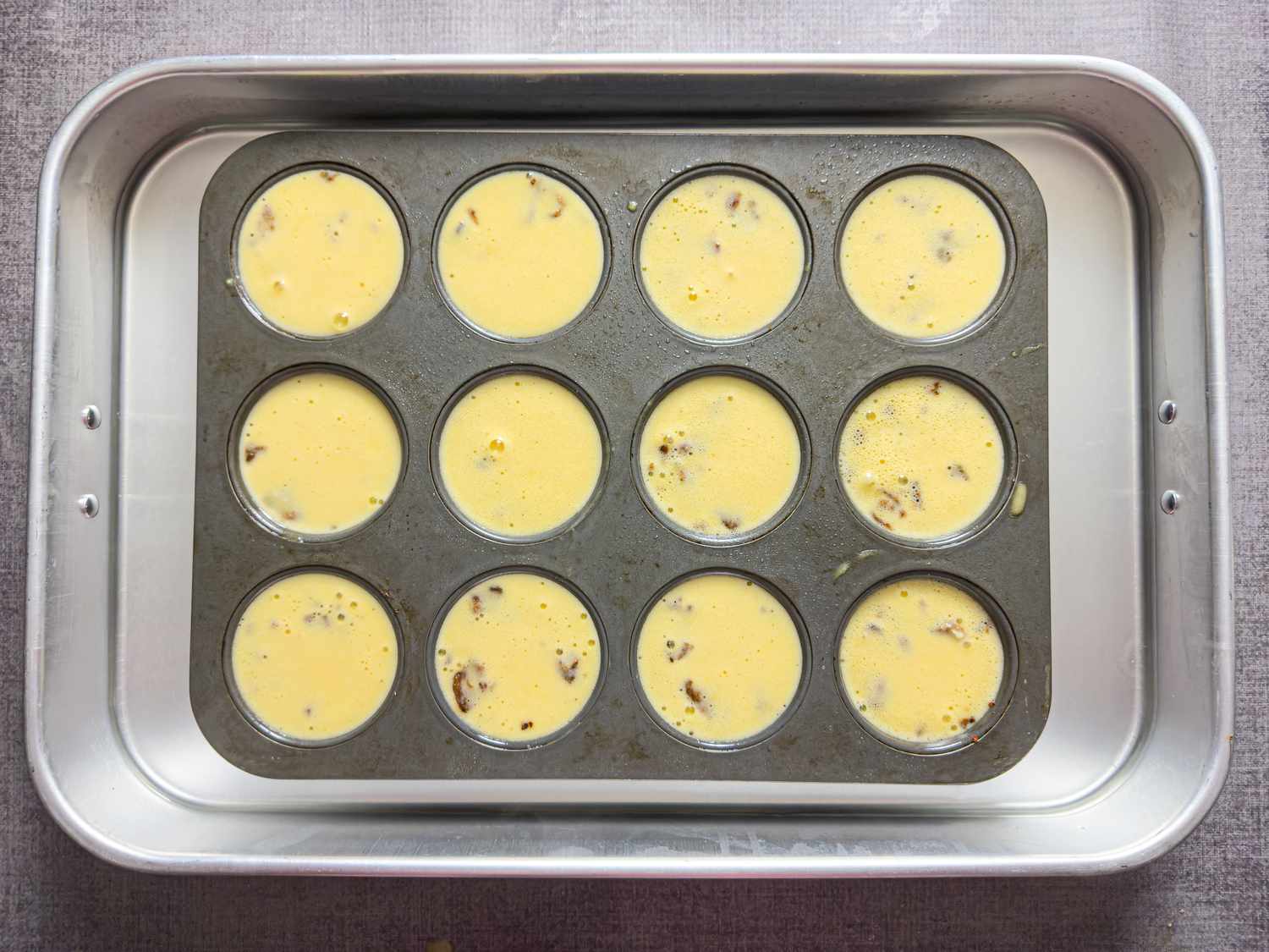 Muffin tin holding twelve portions of raw egg mixture with bits of bacon ready to be baked, placed in a baking tray