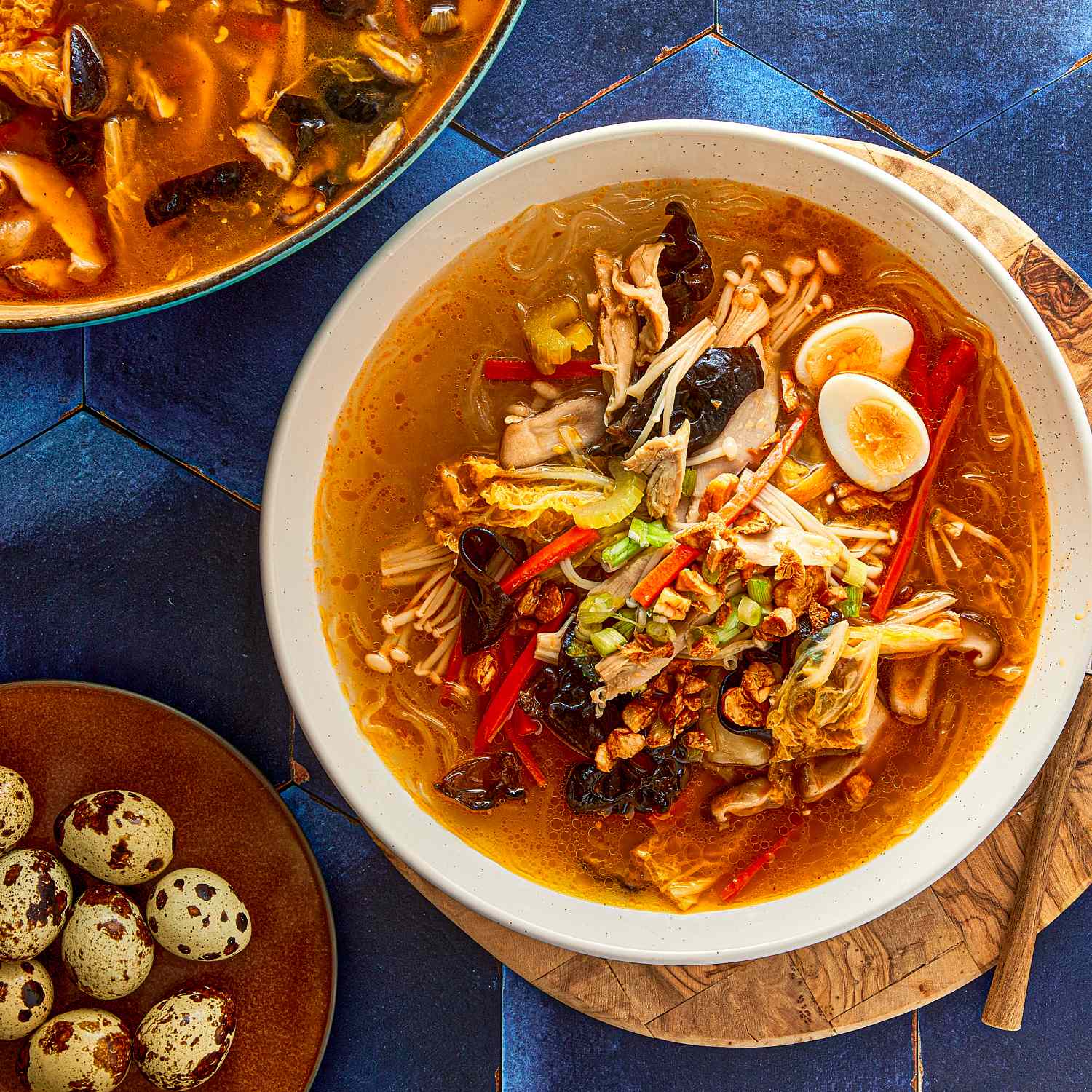 Bowl of chicken sotanghon with garnishes accompanied by a side of quail eggs on a blue surface