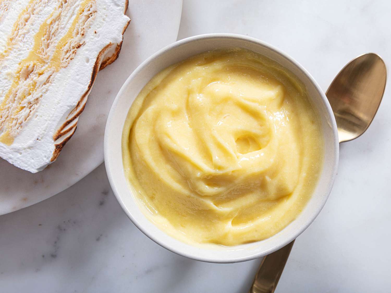Overhead angle of lemon Meringue in a small bowl with a gold spoon. Next to the bowl is a slice of lemon meringue icebox cake 