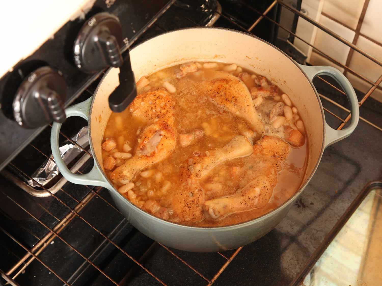 Top down view of a Dutch oven containing simmering beans, chicken, salt pork, and sausages.