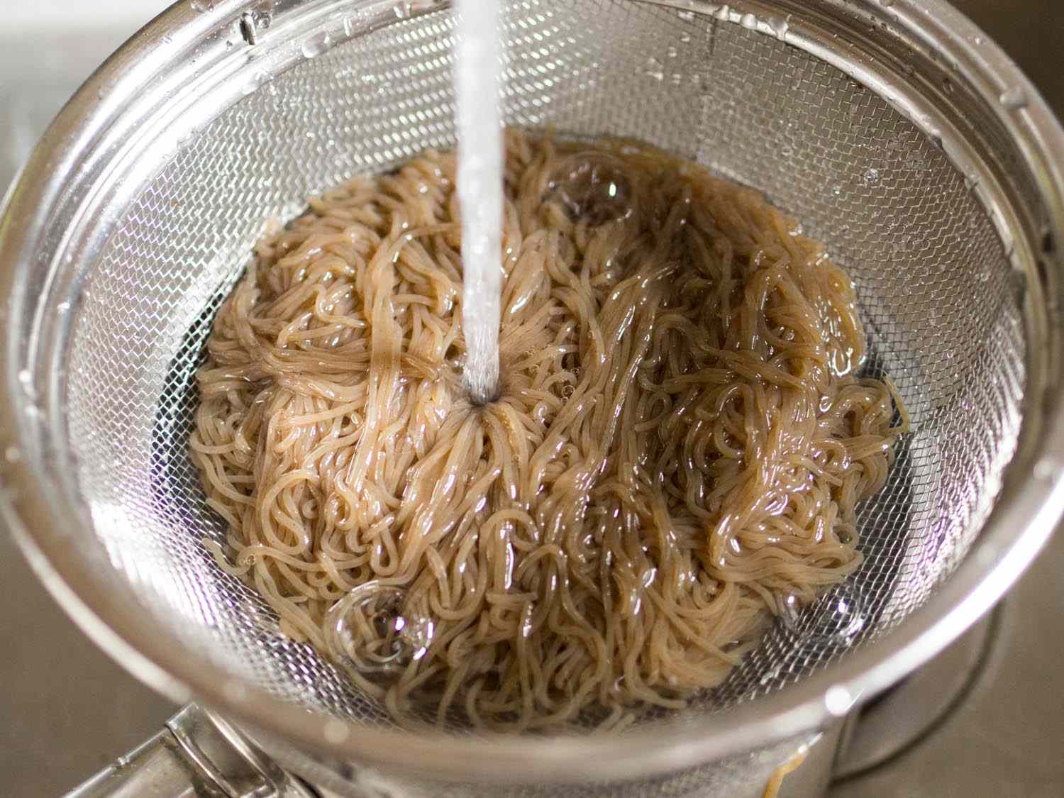 Cooked noodles being drained in a colander and rinsed with running water.