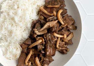 A closeup shot of stir-fried beef with mushrooms and butter. The beef and mushrooms are plated in a round white bowl with white rice on a white tile background, and the shot shows how the mushrooms have been seared, while the feed is tender and in a somewhat glossy sauce.