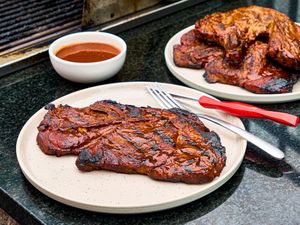 Cooked pork steak on a plate with barbecue sauce and utensils additional steaks in the background on a platter on a black countertop