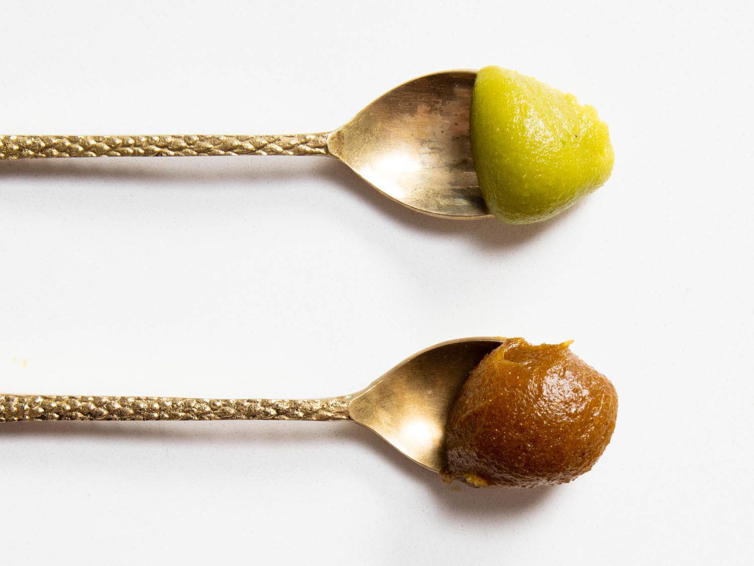 A spoon of raw pistachio paste on top and toasted pistachio paste on bottom. The raw pistachio paste has a green color, while the toasted version is olive. 