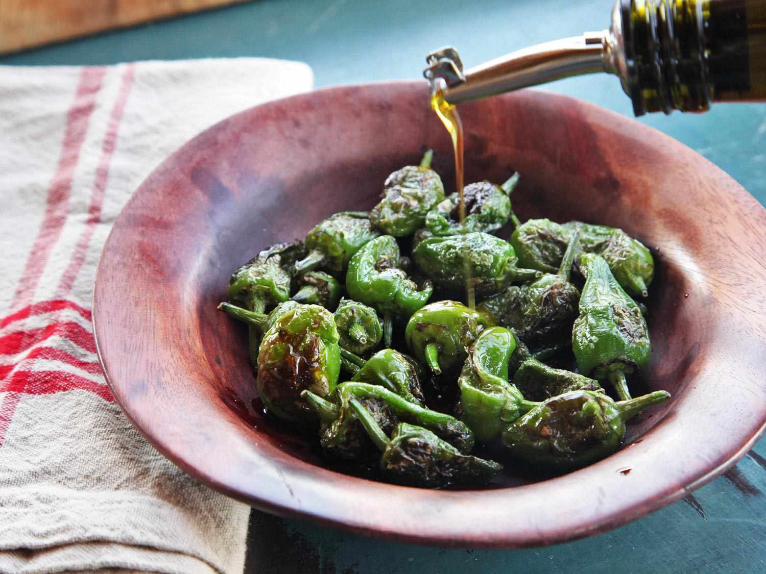 Olive oil pouring over wooden bowl of blistered padrón peppers