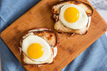 Two Croque Madame sandwiches, topped with sunny-side up eggs on a wooden board on a blue cloth.
