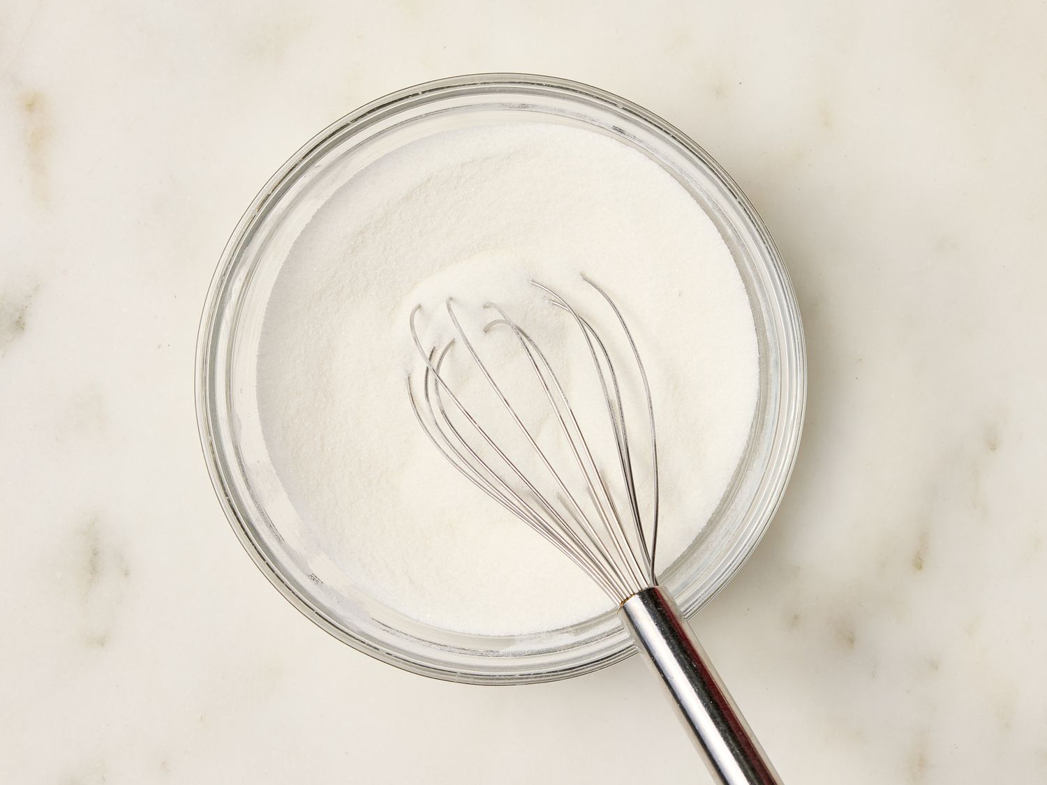 A whisk in a bowl containing powdered ingredients, placed on a marble surface
