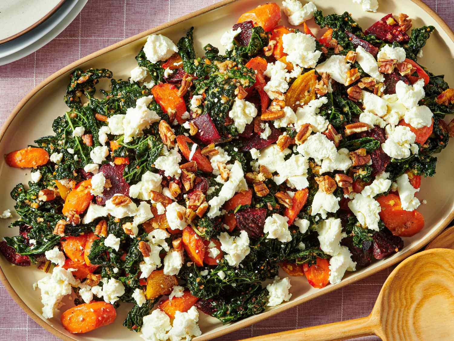 A salad made with kale roasted root vegetables and crumbled cheese topped with nuts on a rectangular serving dish