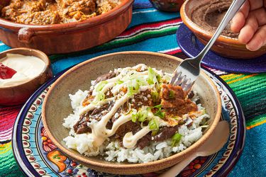 Dish of morisqueta on white rice, with a fork poking into dish. Dishes of white rice, water, and platter of moriqueta on the side. Table cloth is colorful Mexican striped textile