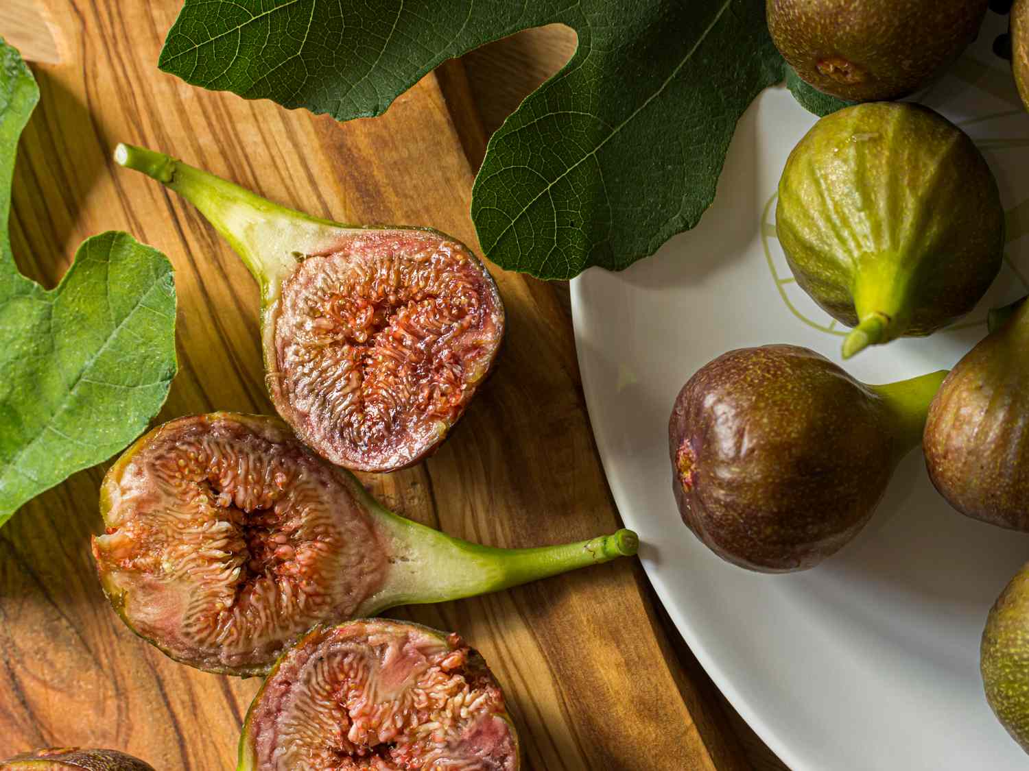 Several fresh figs and leaves some cut open to show their inside placed on a wooden surface and a plate